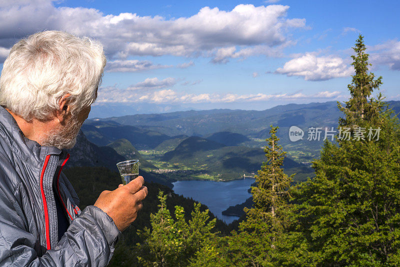 在斯洛文尼亚的朱利安阿尔卑斯山脉的Komna高山小屋后，一名男子喝着饮料，欣赏博欣湖的景色