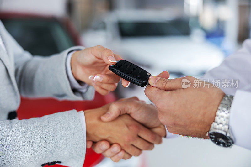 特写的汽车销售商和一个买家握手，而站在汽车沙龙。卖家把车钥匙交给买家。