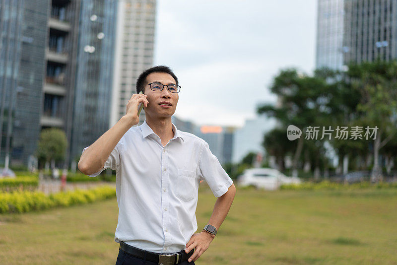 一名亚洲男性白领在办公楼外的草地上用手机打电话