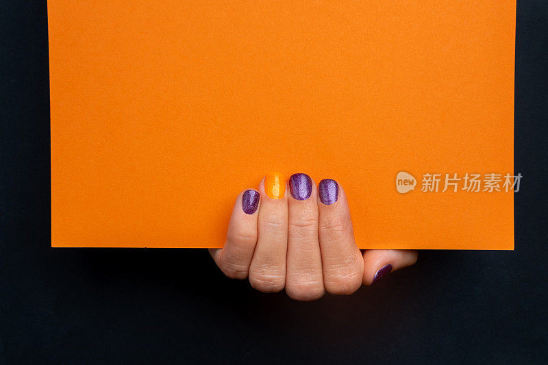 手拿紫色凝胶指甲油，黑色背景上的空橙色纸，万圣节邀请，上面的文字。