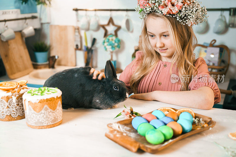 美丽的女孩头戴花圈庆祝复活节。烹饪蛋糕，在厨房桌子上画鸡蛋。节日快乐在一起。和复活节兔子玩，兔子