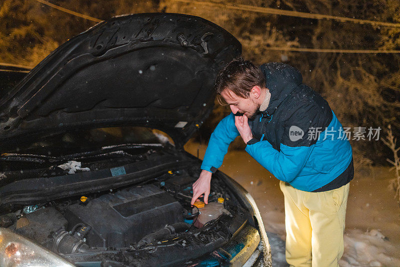 一个年轻人，被困在雪地里，一边打电话一边看他的汽车引擎盖下