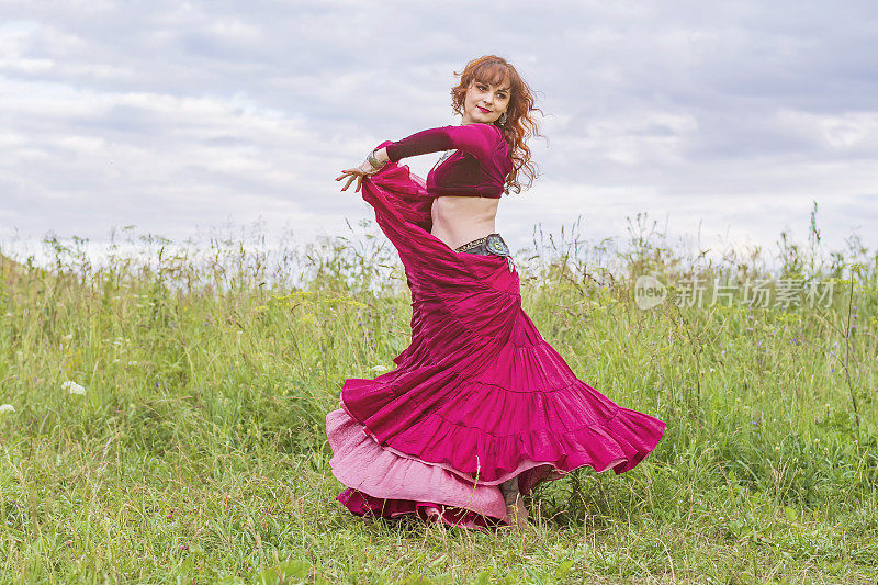 那个红头发、穿红裙子的漂亮姑娘正在绿色的草地上跳民族舞蹈。