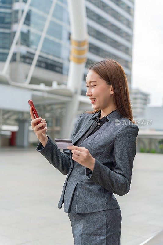 一名年轻的亚洲女商人穿着灰色的办公套装，在商业区用她的智能手机使用信用卡进行在线交易，她微笑着自信