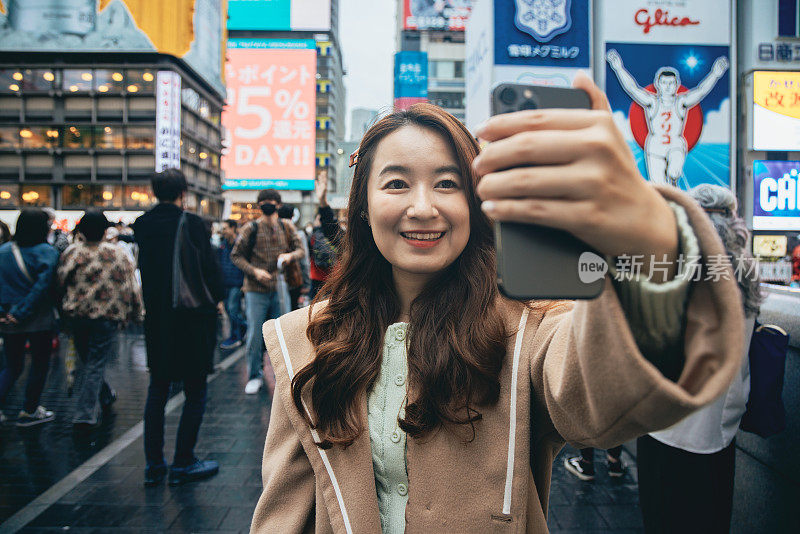 在大阪道顿堀，一位亚洲女性游客正在用手机自拍。她捕捉了她的旅行记忆和直播，并在社交媒体上与朋友和家人分享了她的照片。她正在分享当地的美景。