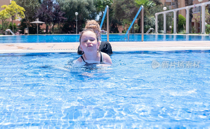 一位年轻的白人妇女和一个8岁的女孩正在室外游泳池里放松地游泳。