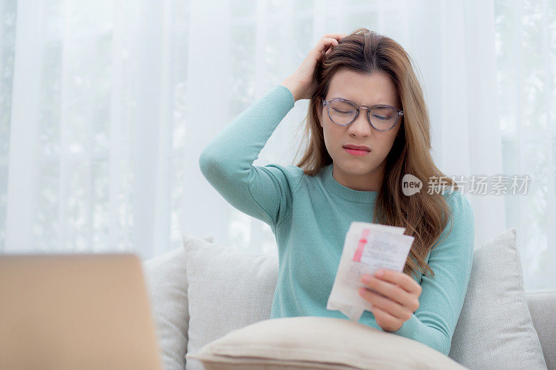 年轻的亚洲女人戴着眼镜，坐在沙发上担心家里的财务账单和压力，不快乐的女人对逾期的贷款或税收、生意和生活方式感到沮丧和焦虑。