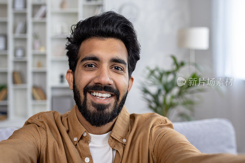 发型时髦的印度男性在光线充足的公寓室内自拍，双手拿着相机。有魅力的没刮胡子的家伙微笑着为简历拍照