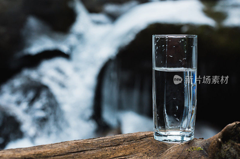 木头上放着一个透明的玻璃玻璃杯，里面装着可以饮用的山泉水，背景是一条清澈的山间河流，在冬天湍急的水流