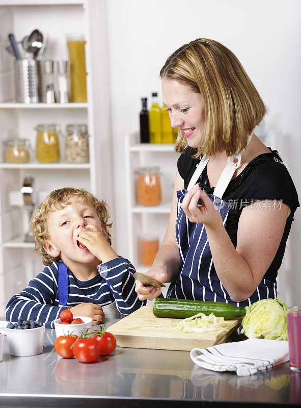 小男孩在吃水果，他的妈妈在做饭