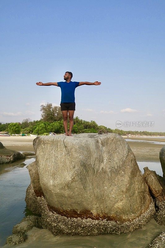 印度男子练习山姿势伸出手(山式伸出手)瑜伽姿势在岩石，水的边缘，帕洛伦海滩，果阿，印度