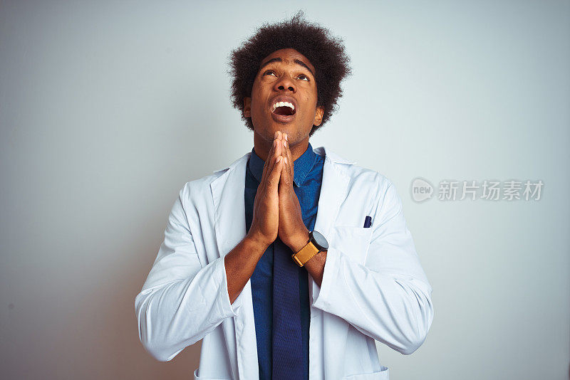 年轻的非裔美国医生男子穿着大衣，站在孤立的白色背景乞求和祈祷的手在一起，脸上的希望的表情非常情绪化和担心。请求宽恕。宗教的概念。