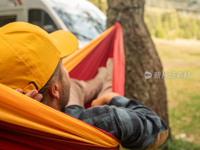 一名男子在吊床上休息，远处是露营车