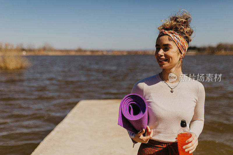 前面的观点微笑的年轻女子与一瓶果汁和一个瑜伽垫在前面的湖