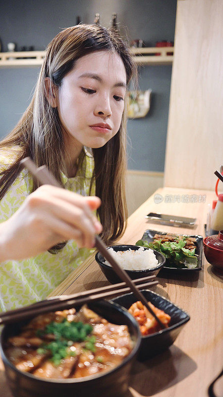 年轻一代Z在一家日本餐厅的木桌上，面带微笑的亚洲女性吃着烤猪肉米饭和鸡蛋。