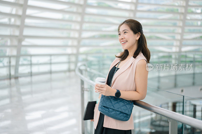 亚洲妇女望着远方，微笑着拿着咖啡，享受工作日的城市办公大楼
