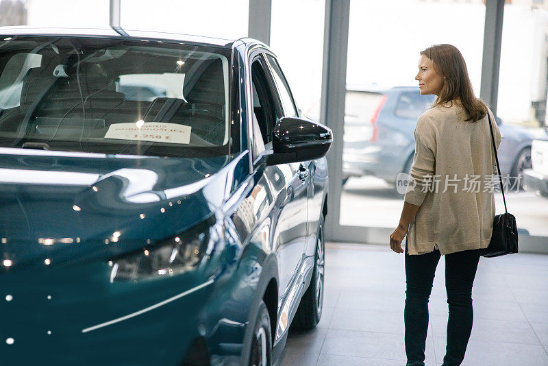 一名成年女子在一家汽车经销商的室内观看一辆新的SUV汽车，她站在汽车旁边，背景是停在户外的汽车