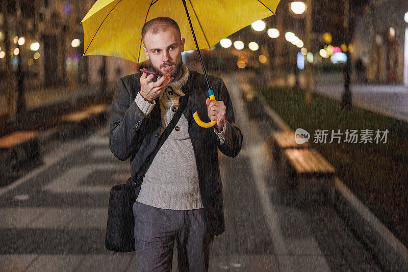 一个英俊的年轻人在雨夜走在街上，打着黄色的雨伞，用手机聊天。