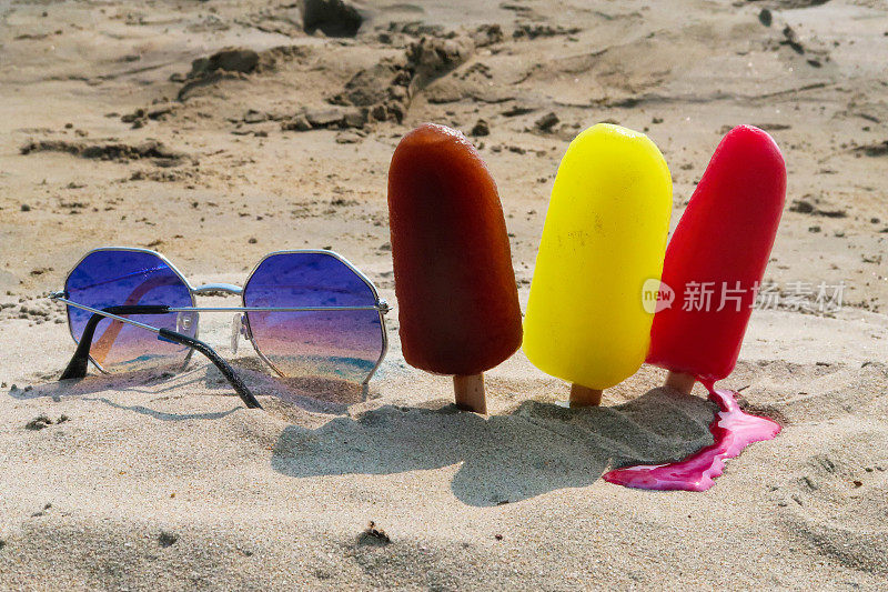 近距离图片，三排冰棒，木制的冰棒棒卡在沙滩上，红色，黄色和棕色的冰棒除了镜像太阳镜，融化的草莓，柠檬和可乐口味的冰棒，重点放在前景