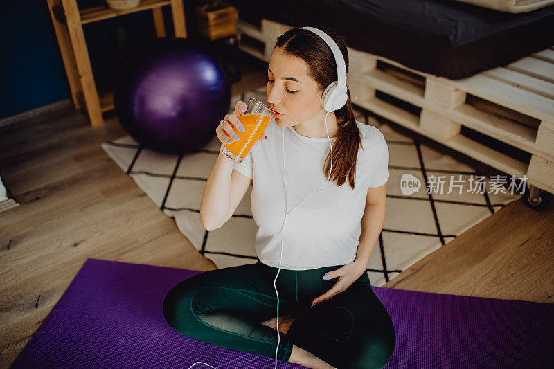 一个年轻的孕妇坐在家里的运动垫上，喝着橙汁