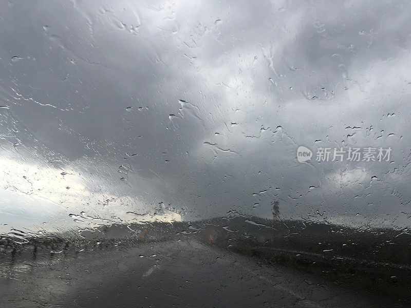 下雨天开车。雨点落在汽车挡风玻璃上，多云的天空和空旷的道路背景