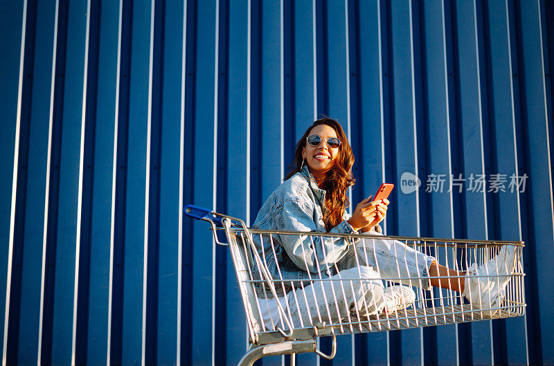 微笑的女人拿着智能手机，推着购物车