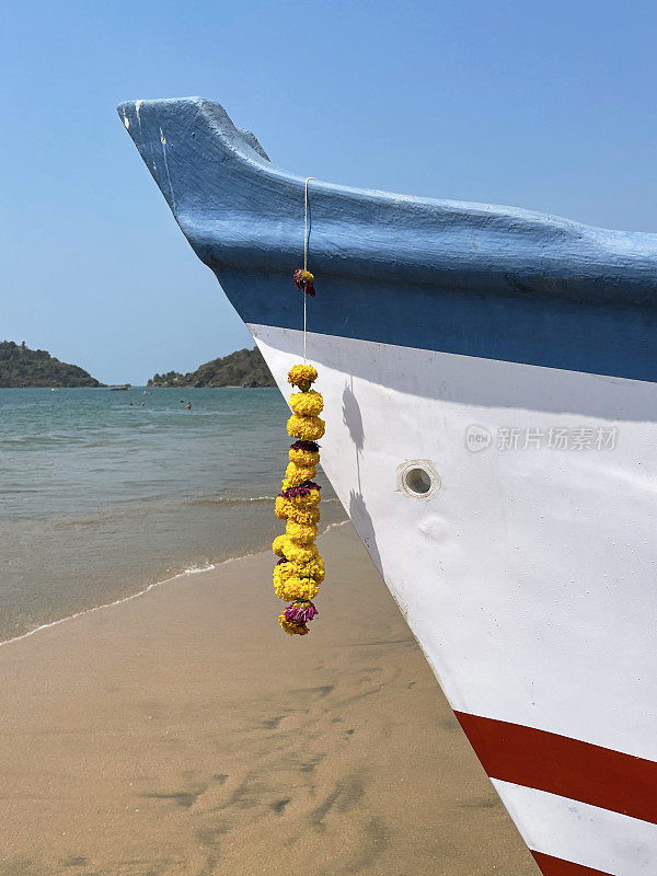停泊在Palolem海滩上的旧木制印度渔船的船头特写图像，船体涂成蓝色，红色，白色，黄色万寿菊，花头，船头上的花环，重点放在前景上