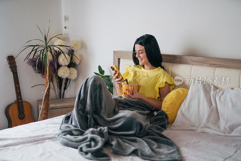 一个年轻的女人正在床上吃早餐，用她的智能手机。