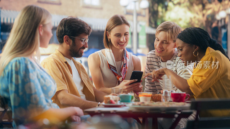 一位年轻女性和多民族朋友坐在露台上，用智能手机展示度假照片。年轻男女聚集在咖啡馆里，进行有趣而有思想的交谈
