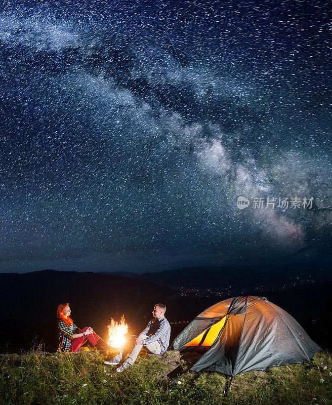晚上露营。幸福的背包客夫妇坐在篝火和帐篷下，在难以置信的美丽星空下。村庄在山谷的背景。长时间曝光
