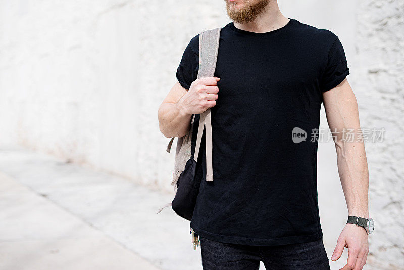 年轻的蓄着胡须的男子穿着黑色t恤，背着背包在外面摆姿势。背景是空白的白色混凝土墙。Hotizontal模型，正面视图