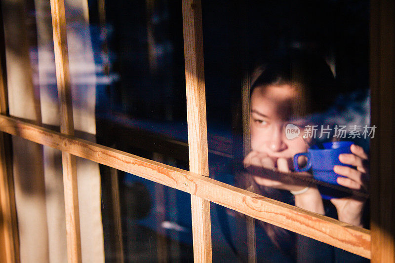 沉思的年轻女子透过窗户看和喝茶