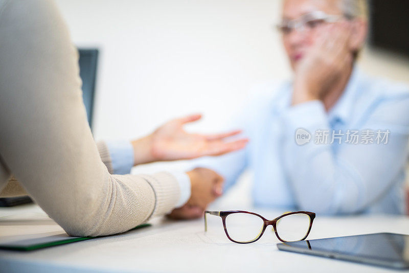 一位年轻女性治疗师的桌子上放着一副眼镜，当时她正在和一位资深女性交谈。