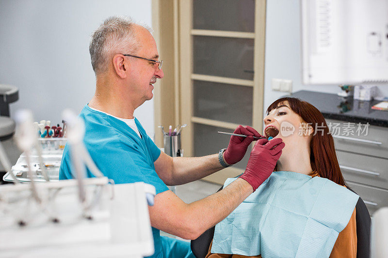 一位年轻女子来看牙医，张着嘴坐在牙科医生的椅子上。资深微笑的男人牙医检查她的牙齿。快乐的病人和牙医的概念。