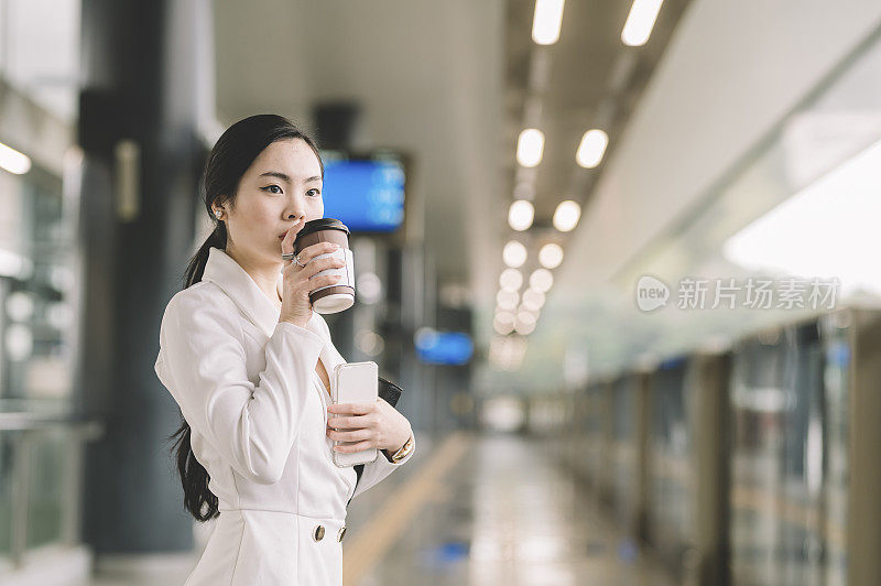 一位年轻的亚洲华裔女商人在单轨火车站边等车边喝咖啡