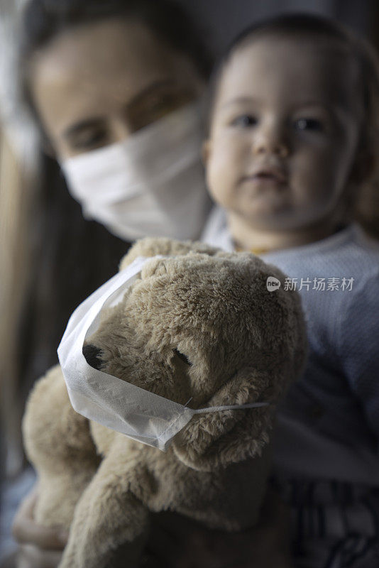 可爱的小女孩和带着防护面具的妈妈和她的泰迪熊站在窗边