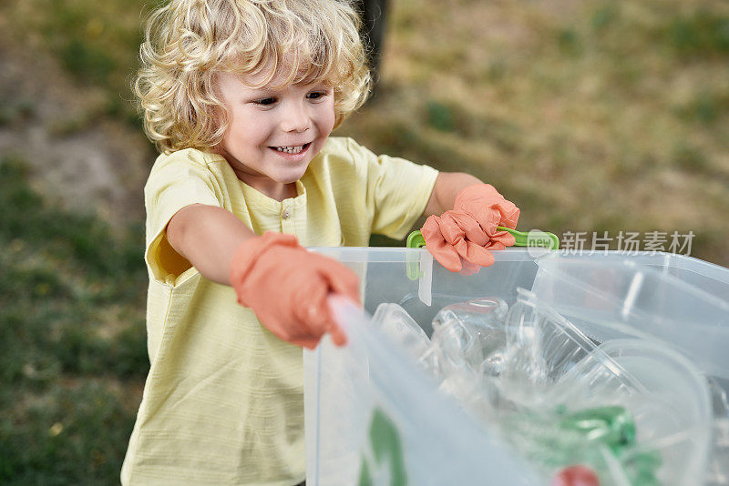 回收和孩子。可爱的小男孩戴着橡胶手套，手拿回收箱，微笑着和父母在森林或公园收集塑料垃圾