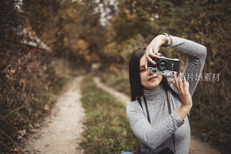 在大自然中摄影——快乐的年轻女子微笑着享受大自然，走过画着秋天颜色的森林，戴着复古的相机拍摄树叶