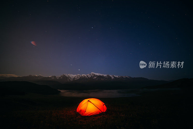 大气的山景观与生动的橙色帐篷在夜晚星空下的小山。橘红色的帐篷在星夜里闪烁着雪山的景色。在大自然的大山中过夜。