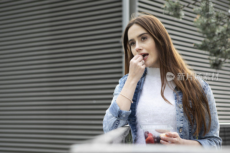 一名身穿花裙、白色套头衫和牛仔夹克的年轻女子站在一座金属建筑旁边，吃着塑料(聚对苯二甲酸乙二醇酯)容器里的水果