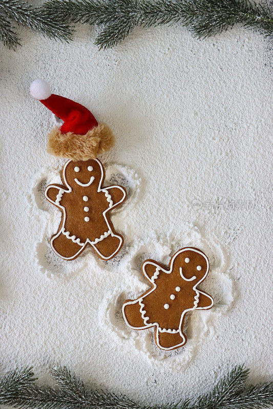 两个自制的姜饼人，用白色皇家糖霜冰镇，一个戴着圣诞老人帽，在糖霜的背景下制作雪天使，云杉针的框架，升高的视角