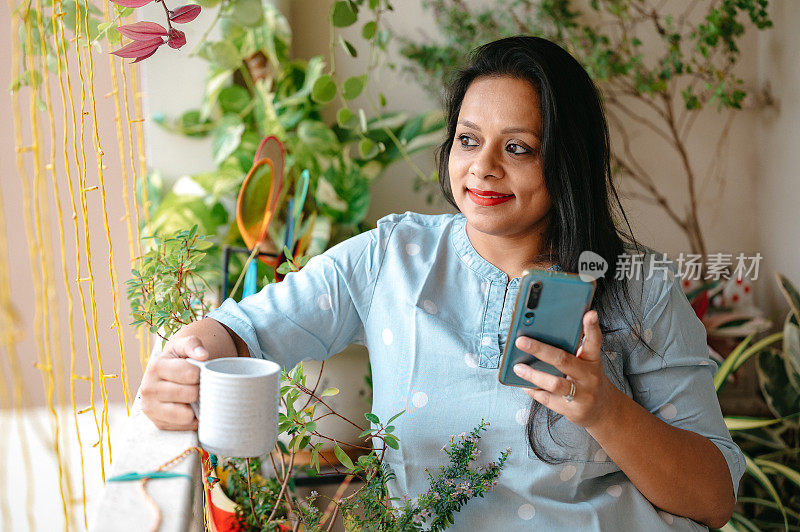 在阳台上用智能手机和拿着一杯茶或咖啡的年轻女子。