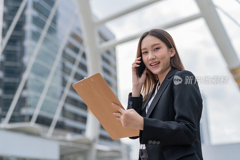 在曼谷市中心的一个商务区，年轻迷人的亚洲职业女商人女士面带微笑，在看完剪贴板上的市场和销售报告后，一边看着相机一边打电话