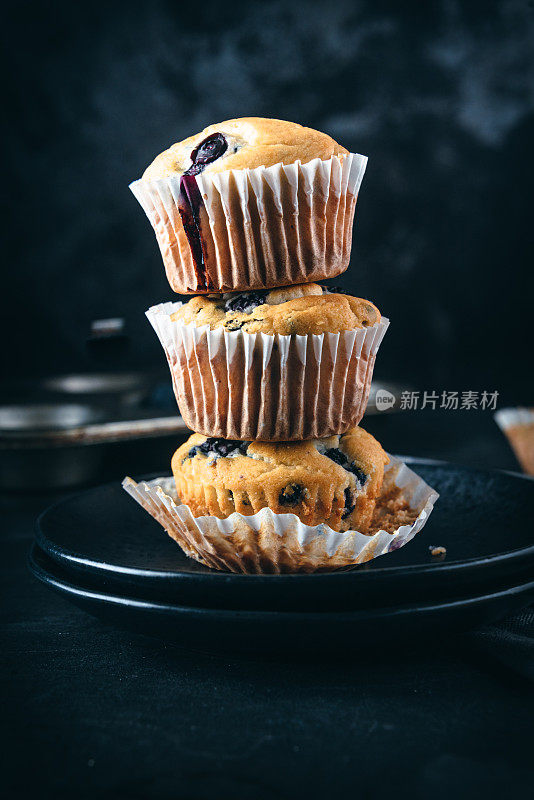 一叠自制的蓝莓松饼装在纸蛋糕盒里，周围是新鲜的蓝莓。