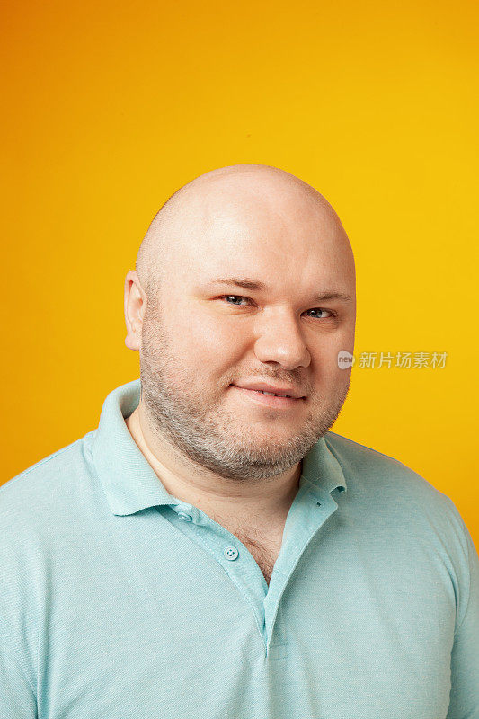 特写工作室肖像超重中年成年白人男子剃了头，穿着蓝色polo衫，反对黄色背景