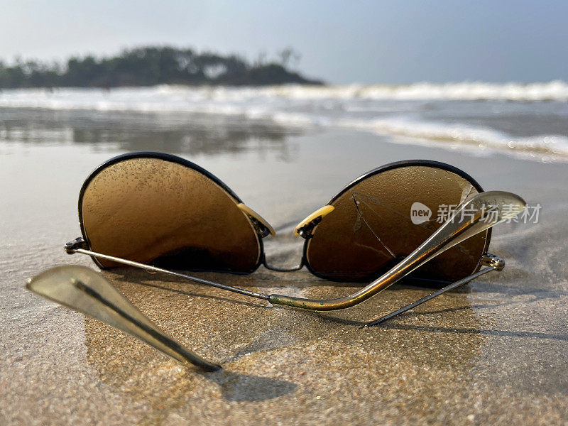 一对飞行员的特写图像，金属框，着色，镜面太阳镜被冲到潮湿的海滩沙滩上，在退潮水域的边缘，破碎的镜头，海浪在海岸上破碎，聚焦在前景
