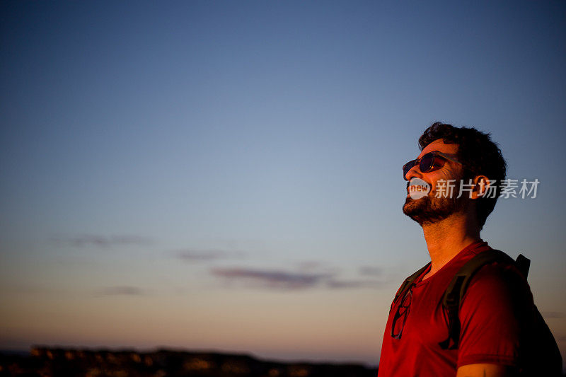 一名身穿红衬衫、背着双肩包的游客微笑着向夕阳下的田野望去