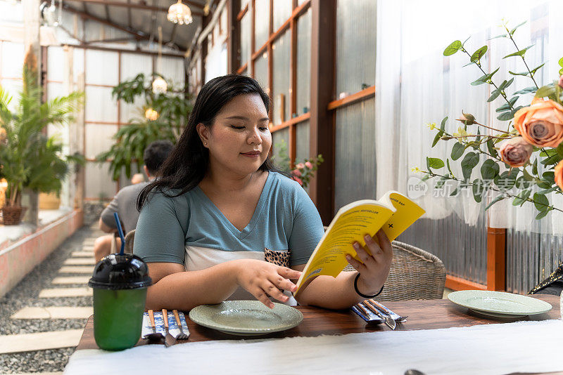快乐的中年亚洲女性喝咖啡，读一本有趣的书，休息一下工作，坐在咖啡馆，周末放松的时间。良好的心理健康和幸福感。