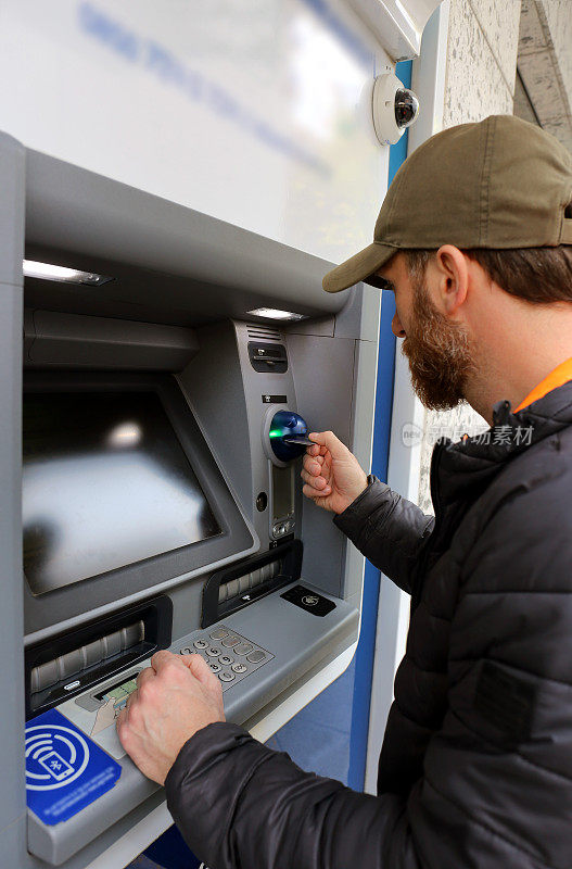 一名蓄着胡子的银行客户使用借记卡和信用卡通过自动柜员机完成交易的垂直照片
