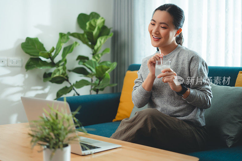 亚洲妇女坐在客厅的沙发上用吸管喝凉水休息。